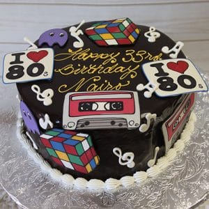 BD-063 Ganache with 80’s theme/music notes: A chocolate cake topped with Rubik’s cubes, cassette tapes, I Love 80s signs, purple Pac-Man ghosts, and music notes. Yellow writing on top reads “Happy 33rd Birthday Naiso.”.