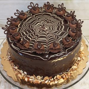 The BD-016 Boston Cream cake is a round chocolate cake with a glossy glaze, topped with a white and dark chocolate spiderweb, swirls of chocolate cream, chocolate decorations, and sliced almonds around the base.