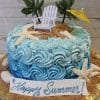The BD-137 is a beach-themed cake featuring blue swirled frosting, white starfish, edible sand, palm trees, a mini white chair and yellow umbrella, plus a “Happy Summer!” sign on the front.