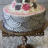 BR-013 is a round white cake with gray and pink roses on top, gray scalloped icing on the sides, and pink writing. It's displayed on an ornate gold stand with crystal accents against a white textured background.