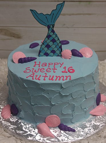 The CB-021D is a blue birthday cake decorated with pink and purple seashells, a mermaid tail topper, and "Happy Sweet 16 Autumn" written in pink icing. The cake sits on a silver board.