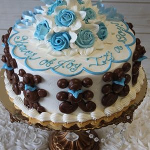 BB-006 is a round white cake topped with chocolate teddy bears, blue and white icing flowers, and blue "Shower of Joy, It’s a Boy!" lettering, displayed on a gold crystal stand.