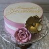 The BD-060A Champagne Pink/Gold Flowers cake is a round pink and white birthday cake with “Happy Birthday” in gold icing, topped with a large pink fondant rose and gold flower, shown on a silver cake stand.