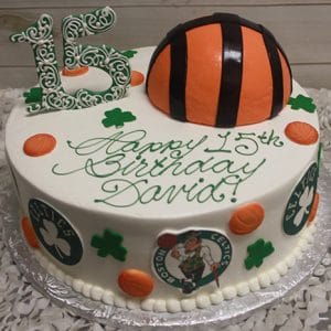 The ST-005 Celtics Basketball/Number cake features a basketball half on top, green shamrocks, orange circles, Celtics logos, and a large green “15.” “Happy 15th Birthday David!” is written in green icing.