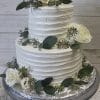 TC-003 is a two-tiered white cake with textured frosting, adorned with white roses, greenery, and sprigs, displayed on a silver stand against a light wood background.