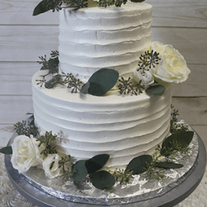 WD-010: Two-tiered white cake adorned with white roses, green leaves, and delicate sprigs, presented on a silver stand against a light wooden background.