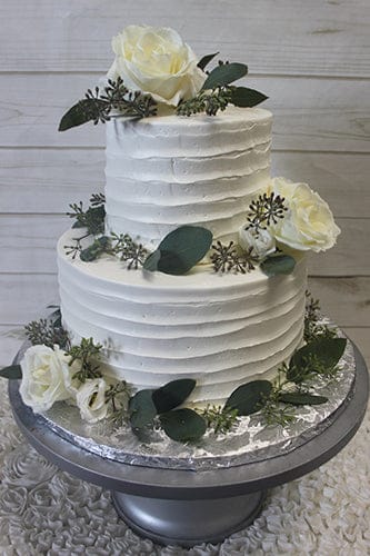 TC-003 is a two-tiered white cake with textured frosting, adorned with white roses, greenery, and sprigs, displayed on a silver stand against a light wood background.