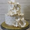 TC-004 is a two-tiered white cake featuring large white flower toppers, gold pearl accents, a quilted pattern with swirls, and sits on a gold board adorned with a rhinestone ribbon at the base.