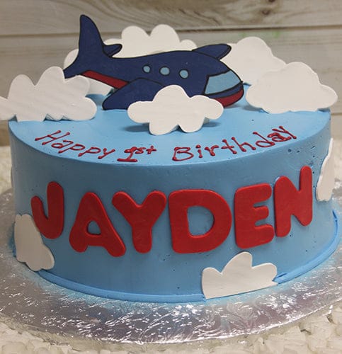 The CB-027A is a blue birthday cake with white clouds, a cartoon airplane topper, large red letters spelling "JAYDEN," and "Happy 1st Birthday" written in red icing.