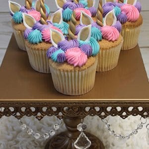 Six unicorn-themed cupcakes with pastel pink, purple, and blue frosting, gold unicorn horns, and white ears are displayed on an ornate gold cake stand with crystal decorations.
