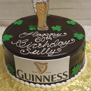 The BD-024G Guinness is a round chocolate cake with green shamrocks, a pint of beer topper, "Happy 60th Birthday Sully," a Guinness logo on the side, and is presented on a gold board.