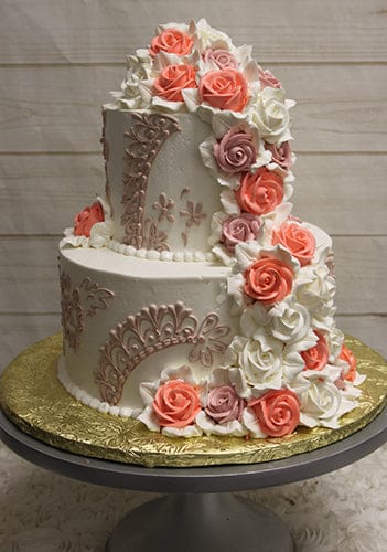 The WC-000E is a two-tiered white cake with lace-like piping and cascading buttercream roses in pink, coral, and white, presented on a gold-covered cake board atop a gray cake stand.