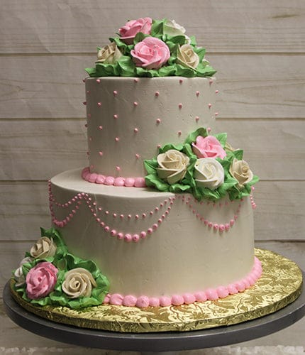 The WC-000H is a two-tiered white cake adorned with pink and white icing roses, green leaves, and pink decorative dots and swags. It is displayed on a round gold board against a light wood background.