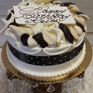 The BR-047 is a round birthday cake adorned with white and dark chocolate seashells, a black ribbon with white dots, and a “Happy Birthday Keith” card in chocolate script, presented on a gold cake stand.