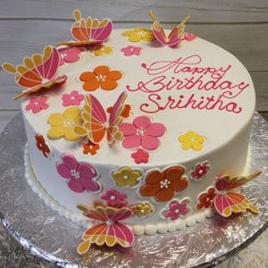 The BD-072 is a round birthday cake topped with colorful fondant butterflies and flowers, “Happy Birthday Srihitha” in pink icing, and presented on a silver board.