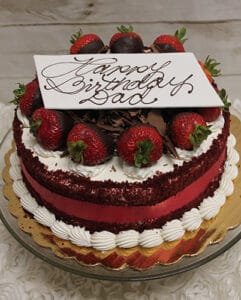 The BD-006A Red Velvet is a round cake topped with strawberries, chocolate shavings, and a white chocolate “Happy Birthday Dad” plaque, presented on a gold cake board and glass stand.