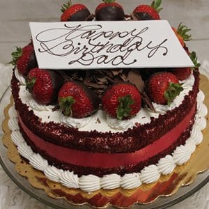 The BD-006A Red Velvet is a round cake topped with strawberries, chocolate shavings, and a white chocolate “Happy Birthday Dad” plaque, presented on a gold cake board and glass stand.