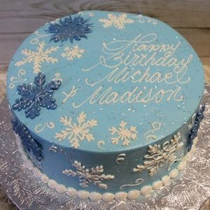 CH-038: A round blue cake decorated with white and blue snowflakes, white pearls around the base, and "Happy Birthday Michael & Madison" in white icing. Displayed on a silver-patterned board.