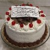 The BD-005 Black Forest Cake is a round birthday cake with white frosting, topped with whipped cream, chocolate shavings, cherries, and a white “Happy Birthday Mike” plaque in cursive. Served on a glass stand.