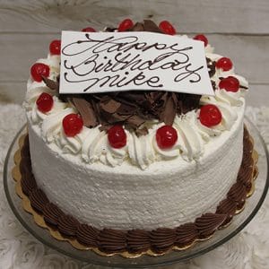 The BD-005 Black Forest Cake is a round birthday cake with white frosting, topped with whipped cream, chocolate shavings, cherries, and a white “Happy Birthday Mike” plaque in cursive. Served on a glass stand.