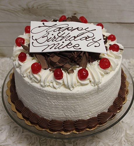 The BD-005 Black Forest Cake is a round birthday cake with white frosting, topped with whipped cream, chocolate shavings, cherries, and a white “Happy Birthday Mike” plaque in cursive. Served on a glass stand.