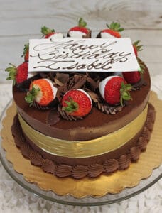 BD-001A Tiramisu decorated with chocolate shavings, chocolate-dipped strawberries, a gold ribbon, and a white card reading "Happy Birthday Isabel" in cursive, displayed on a glass cake stand.
