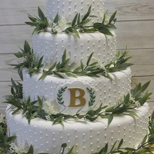 The WD-091 is a four-tiered white cake with dotted icing, green leaves, and small white flowers. A gold letter B inside a wreath decorates the front. Presented on a gold board against a wooden background.