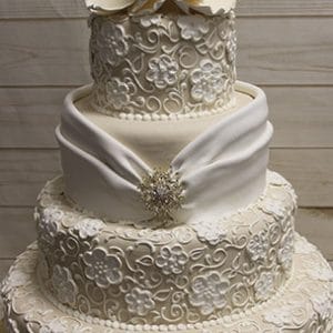 WD-093: Three-tier white wedding cake with intricate floral icing, fondant drape and jeweled brooch on the middle tier, topped with large white flowers. Displayed on a gold base against a wood background.