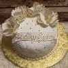 RC-067 is a round white cake with quilted icing, gold pearls, and large white sugar flowers on top. A ribbon with “God Bless Sydney” in gold decorates the cake, which sits on a gold decorative board.