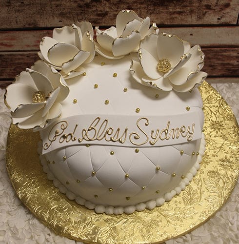 RC-067 is a round white cake with quilted icing, gold pearls, and large white sugar flowers on top. A ribbon with “God Bless Sydney” in gold decorates the cake, which sits on a gold decorative board.