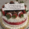 The BD-002A Pina Colada is a round cake decorated with whipped cream, coconut flakes, chocolate-dipped strawberries, chocolate shavings, and a white plaque saying "Happy Birthday Miguel" in cursive, finished with a red ribbon.
