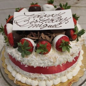 The BD-002A Pina Colada is a round cake decorated with whipped cream, coconut flakes, chocolate-dipped strawberries, chocolate shavings, and a white plaque saying "Happy Birthday Miguel" in cursive, finished with a red ribbon.