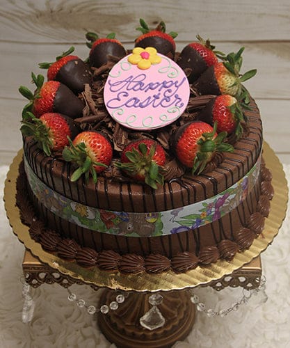 A chocolate cake with chocolate mousse filling, topped with a "Happy Easter" plaque, chocolate-dipped strawberries, and shavings, set on a gold cake stand with crystal accents.