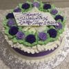 AN-013 is a round white frosted cake adorned with purple and white icing roses, green leaves, a purple ribbon, and a plaque that says "Happy Birthday Kathy Thuy 2-8-2019" in purple icing.