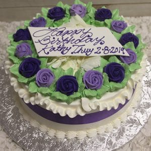 AN-013 is a round white frosted cake adorned with purple and white icing roses, green leaves, a purple ribbon, and a plaque that says "Happy Birthday Kathy Thuy 2-8-2019" in purple icing.