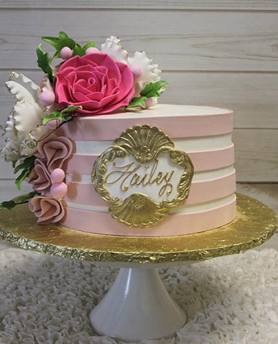 The BR-021 is a round cake with light pink and white stripes, adorned with pink and white fondant flowers, greenery, and a gold ornate frame featuring the name Hailey in elegant script. It sits on a gold board atop a white stand.