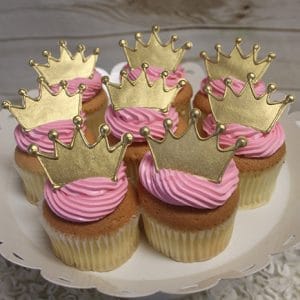 Seven CC-029 cupcakes, chocolate with white chocolate mousse filling, have pink swirled frosting and gold crown toppers, displayed on a white scalloped cake stand against a light wooden background.