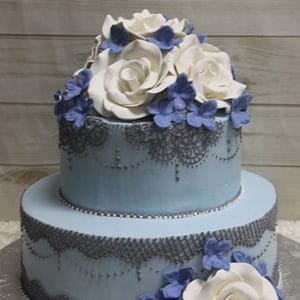 The WD-094 is a two-tiered light blue cake with detailed dark gray icing, topped with large white fondant roses and small blue flowers, displayed on a wooden background.