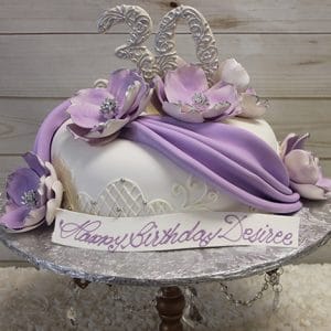 The BD-055A Lavender Swag is an elegant birthday cake adorned with large purple and white flowers, lavender fondant drapes, a “30” topper, and “Happy Birthday Desiree” written on the front, presented on a silver stand.