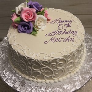 The BD-060D cake features pale pink, lavender, and white icing roses with ornate swirl icing. "Happy 50th Birthday Mei An" is written in purple on top. The round white cake sits on a decorative silver base.