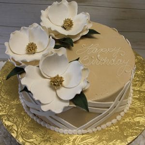 BR-036 White Magnolias: Round birthday cake with beige frosting, "Happy Birthday" in white script, topped with three large white sugar magnolias and green leaves, presented on a gold-patterned board.