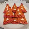 Eight CE-001a Birthday cookies shaped like orange party hats with pink decorations and white number 1 icing are arranged on a white scalloped plate.
