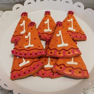 Eight CE-001a Birthday cookies shaped like orange party hats with pink decorations and white number 1 icing are arranged on a white scalloped plate.