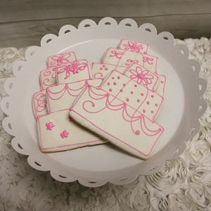 CE-003b bridal wedding cake-shaped cookies, adorned with pink icing, are beautifully arranged on a white scalloped-edge plate set on a textured white surface.