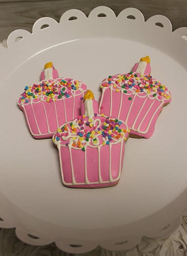 The CE-001b Birthday features three pink cupcake-shaped cookies topped with white icing, colorful sprinkles, and a small yellow candle, all arranged on a white decorative plate.