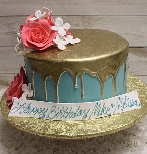 The BD-057B Gold Drip Aqua is a round cake with blue icing, gold drip topping, and large pink and white flowers, displayed on a gold foil board. A banner reads “Happy Birthday Mike & Melina” in blue script.