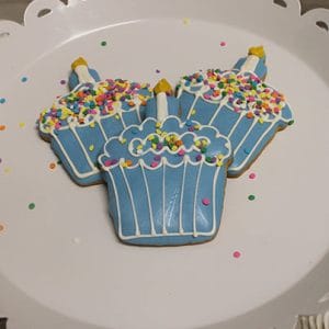 Three CE-001c birthday cupcake-shaped cookies with blue icing, colorful sprinkles, and yellow “candles” on top, displayed on a white scalloped cake stand with confetti scattered around.