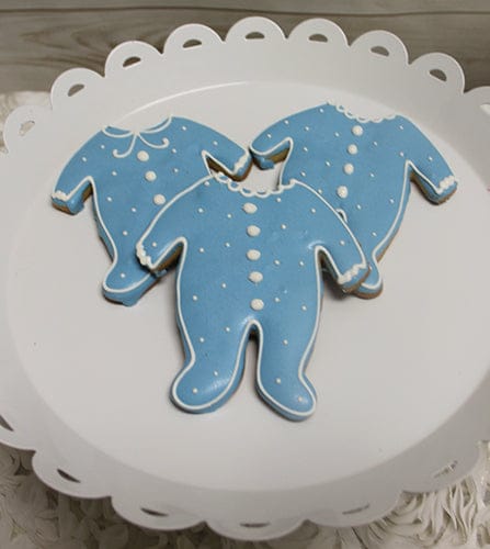 Three CE-004f baby shower cookies shaped like onesies, with blue icing, white polka dots, and collar details, arranged on a white scalloped-edge plate.