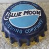 The BD-025 Blue Moon is a round cake styled as a blue bottle cap, showcasing the Blue Moon Brewing Company logo and “Established 1995” on top, presented on a gold base with a textured white background.