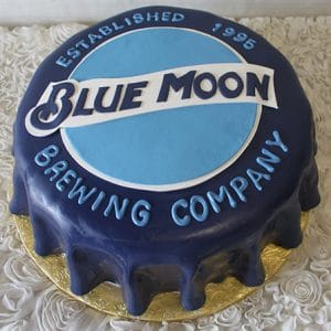 The BD-025 Blue Moon is a round cake styled as a blue bottle cap, showcasing the Blue Moon Brewing Company logo and “Established 1995” on top, presented on a gold base with a textured white background.